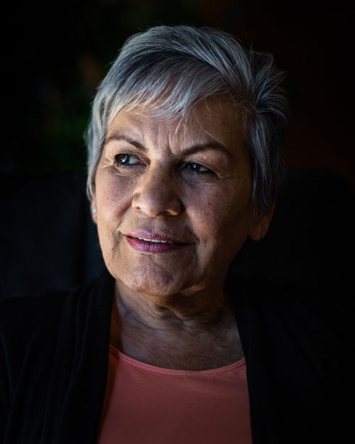 Older woman with short gray hair is smiling in a cloud of black. She is wearing a pink shirt and is looking into the distance where a light is cominf from. This pictures is being used as the feature image for Senior Scholars program.
