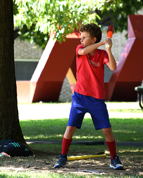 Picture of an child in a red shirt and blue shirt holding a baseball bat under a tree.