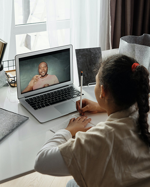 Small girl wiritng with a pencil at a desk in front of a laptop with a man speaking on screen.