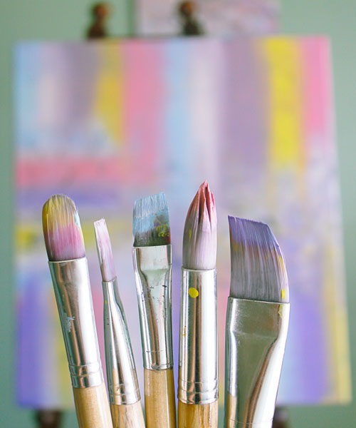 Picture of paint brushes which have variety of pastel colors on them. The bruses hace a painted canvs in the background with yellow, purple, pink and blue lines.